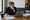 Person writes notes beside an open laptop, papers, and coffee mug at a wooden desk.