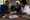 Three adults review documents together at a table, with a cross visible on the wall behind them.