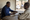 Two men sit at a wooden table indoors, writing and speaking while people gather outside the open doorway.