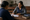 Two women sit at a wooden table as one writes on paperwork inside a church.