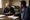 Two people sit at a wooden table in a church, conversing as one writes in a notebook.