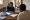 Woman points to a document while speaking with a seated person at a table; a health worker walks in the background.