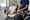 A healthcare professional writes on a clipboard while a seated person waits beside sports equipment.