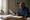 A man sits at a desk reviewing printed documents beside notebooks and a coffee mug.