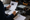 Person writes in a spiral notebook beside printed documents, a stereo unit, speaker, and electronic components.