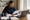 A woman reviews paperwork and writes in a notebook beside an open laptop at a desk.