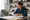 Woman reviews a document beside an open laptop at a desk in a kitchen workspace.