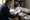 A man reviews printed documents at a wooden desk surrounded by papers, binders, and a coffee mug.