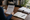 Person reviews a printed document at a desk beside an open notebook, pen, books, and office shelves.