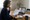Woman reviews a printed document during a video consultation on a laptop at a home dining table.