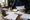 Person writes in a notebook while holding a document at a desk with papers, laptop, and mug.