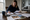 Woman writes on paperwork while reviewing documents, house photos, and a calculator at a dining table.