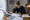 Woman reviews documents and writes at a dining table beside an open laptop, coffee mug, and folder.