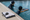 Two swimmers practice in a pool, one holding a kickboard, beside a clipboard and stopwatch on deck.