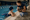 Adult kneels beside an indoor pool, speaking with a child in the water while holding a clipboard.