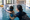 An adult supports a smaller swimmer practicing on their stomach in an indoor pool.