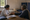 A woman examines a seated man's hand across a desk in a sparsely furnished room.