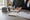 Two people review paperwork at a table, with one writing beside a house model and architectural plans.
