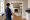 Person holding a clipboard surveys a room with a ladder, fireplace, and protective floor covering.