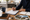 Person writes on paperwork at a kitchen table beside property photographs, a notebook, keys, and a mug.