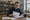 Man reviews paperwork at a wooden counter in a shop stocked with shelves of jars and boxes.