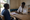 Healthcare worker writes notes while speaking with a seated man across a wooden table in a clinic.