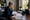 Gray-haired woman reviews documents and writes at a desk beside a window, lamp, books, and potted plant.