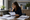Woman wearing glasses reviews a document at a desk surrounded by paperwork, books, and plants.