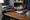 Healthcare worker records notes while examining a patient’s arm across a wooden desk in a clinic room.
