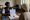 A woman shows an illustrated document to another person across a table in a simple room.