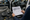 Person holds a vehicle diagram beside an open car, with speakers, amplifier, wiring, and tools on the workbench.
