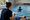 A woman takes notes beside an indoor pool while a swimmer practices in the water.