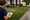 Woman writes on a clipboard while inspecting a sprinkler-watering lawn beside a suburban house and sidewalk.
