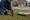 Person kneels on a lawn, hammering a stake into the ground beside sign panels as sprinklers spray.