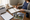 Person writes in a notebook at a desk surrounded by a calendar, teddy bear, house photos, and coffee.