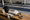 Fencing mask, jacket, gloves, and sword rest on a bench while two fencers practice in the background.