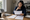 Woman holds and reviews printed documents beside an open laptop at a wooden desk.