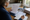 Gray-haired woman wearing glasses reviews paperwork and writes in a notebook at a desk beside house photographs.