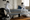 Two fencers in white protective gear practice with swords in a gym, while fencing equipment rests on a nearby bench.