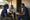 Two people sit with a healthcare worker reviewing paperwork outside a modest building.