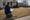 A person kneels on a suburban lawn, measuring the ground with a tape measure and clipboard.