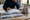 Person writes on architectural plans beside a laptop, house photographs, and a small model home.