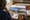 Person reviews home photographs and architectural plans at a wooden desk beside a notebook and coffee.