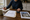 Person writes on a notepad beside a calculator, paperwork, and a house photograph on a wooden table.