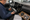 A man holds an electronic component beside an exposed car dashboard, with wiring, a speaker, and paperwork nearby.