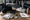 Fencing gear, uniforms, and a notebook sit on a table as people practice in the background.