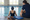 A swimmer rests in the pool beside an instructor seated at the edge, with a kickboard and goggles nearby.