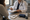 Clinician in white coat writes on a clipboard while a seated person rests their hand near their face.