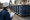 Person in a hard hat holds a clipboard beside a debris-filled roll-off container at a construction site.