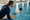 An instructor leans over an indoor pool edge, gesturing toward a swimmer wearing a swim cap.